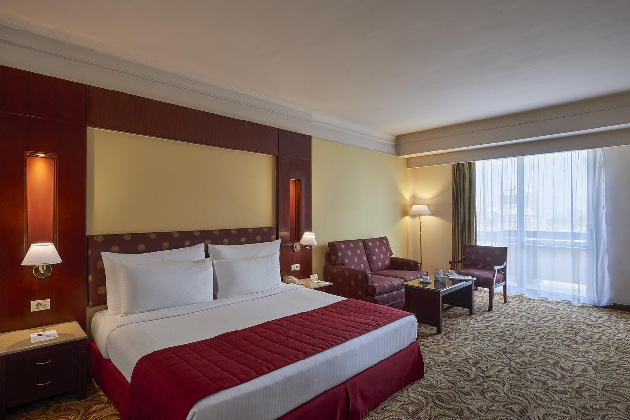 Safir Hotel Cairo Giza Exterior foto The image showcases a hotel room featuring a large bed with white linens and a red bedspread. Behind the bed, there is a decorative headboard. The room includes two armchairs and a small coffee table, creating a seating area. Natural light enters thr