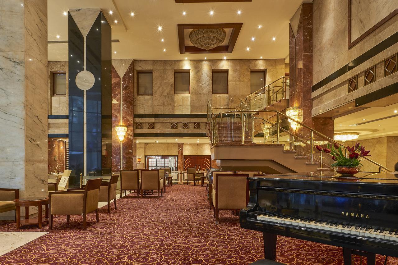Safir Hotel Cairo Giza Exterior foto The image shows an elegant hotel lobby. The space features marble walls and a plush carpet, giving it a luxurious feel. There are seating areas with comfortable chairs arranged around tables. A grand staircase leads to an upper level, and large windo