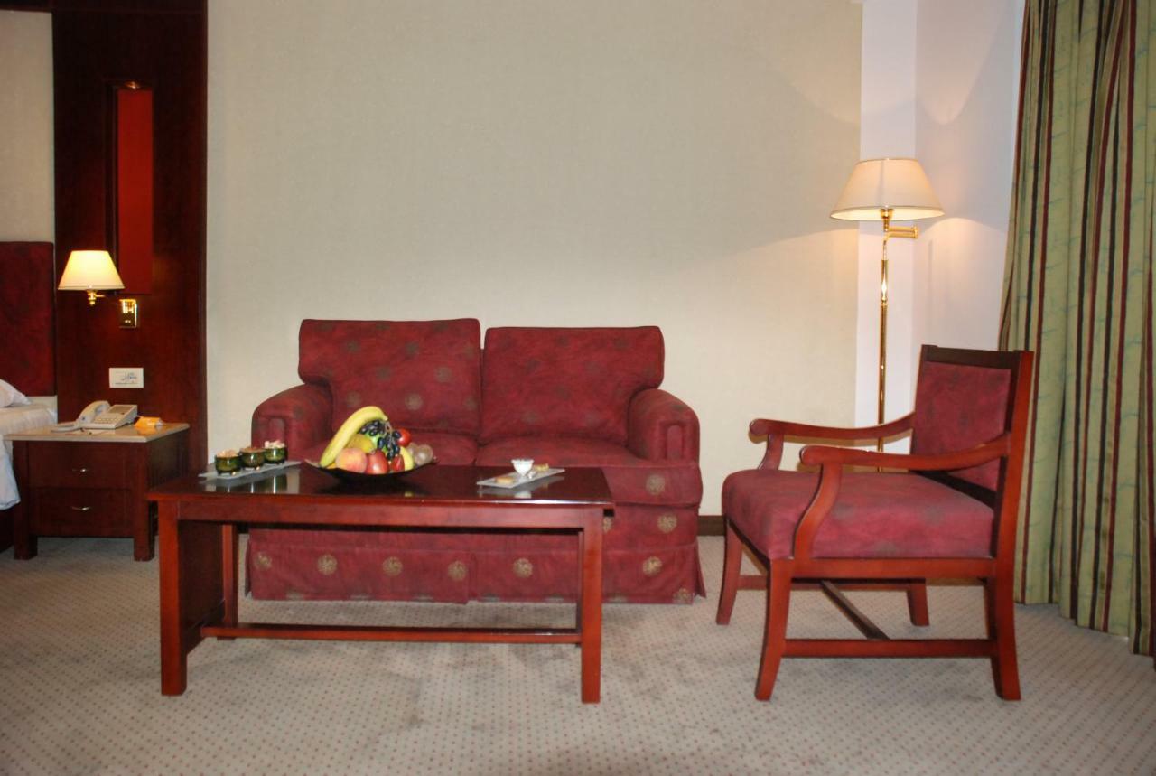 Safir Hotel Cairo Giza Exterior foto The photo shows a cozy seating area in a hotel room. There is a red patterned sofa and a matching armchair placed around a wooden coffee table. On the table, there are some snacks or fruits displayed. A floor lamp is positioned beside the sofa, and t