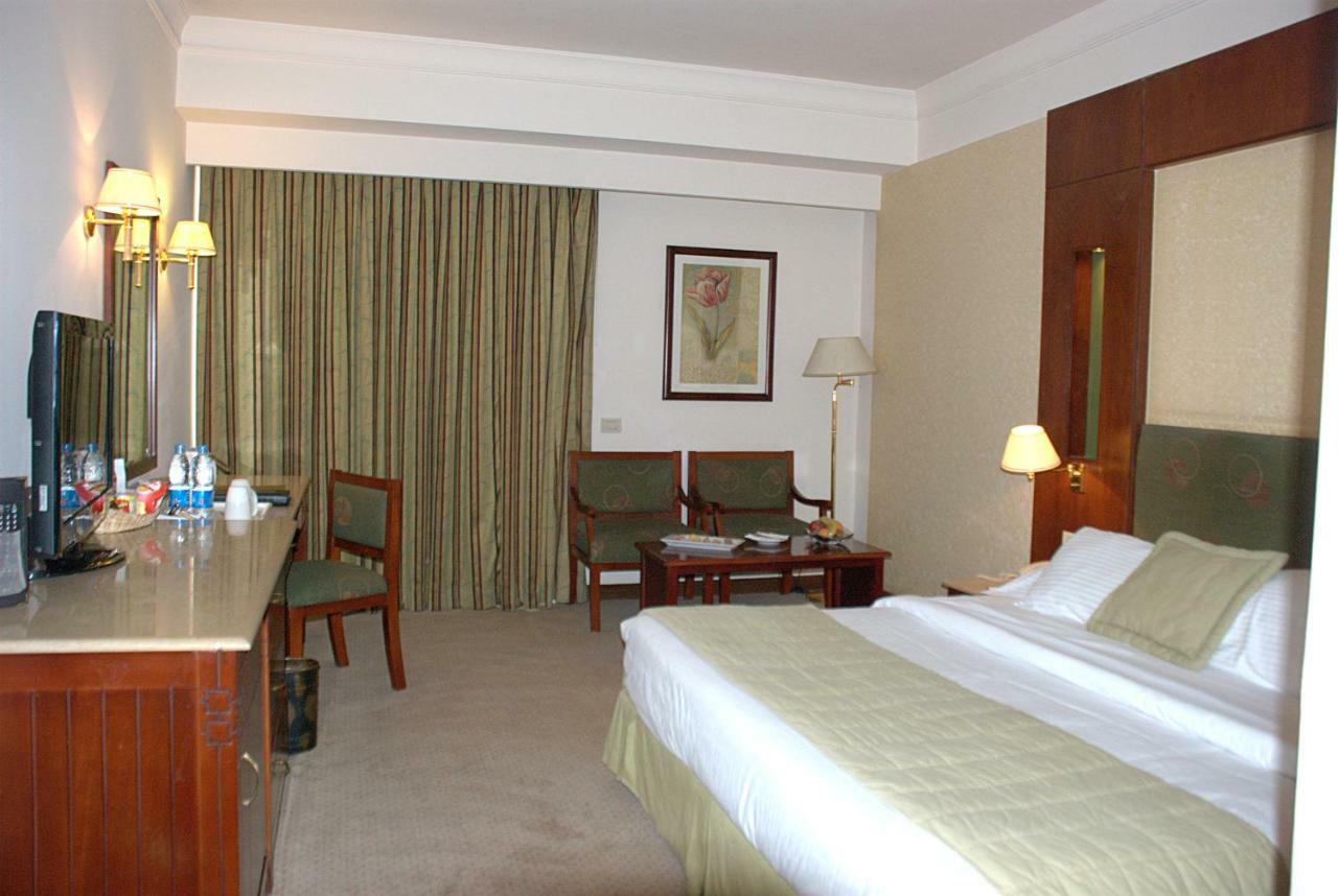 Safir Hotel Cairo Giza Zimmer foto The photo shows a hotel room. It features a bed with white linens and a green throw. On one side of the bed, there's a small bedside lamp. Opposite the bed is a desk with a mirror above it, alongside a couple of chairs and a small table. The room has