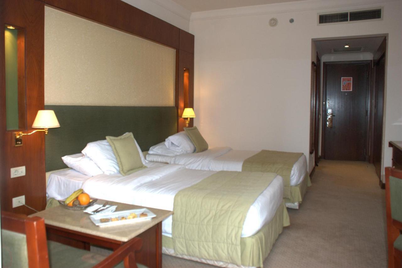 Safir Hotel Cairo Giza Zimmer foto The photo shows a hotel room featuring two single beds with light green bedspreads and white linens. There are bedside lamps on either side of the beds, and the headboard has a soft, textured design. A small seating area is visible with a wooden chai